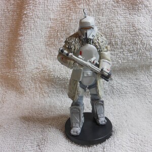 May include: A detailed figurine of a character in a white and gray armored suit, holding a rifle. The figure has a helmet and a fur-like collar. It stands on a black circular base, showcasing a sci-fi or fantasy theme.