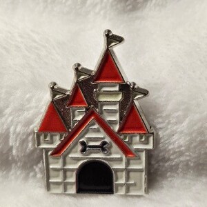 May include: A silver and red enamel pin shaped like a dog house with a bone on the door. The dog house has a white brick pattern and a red roof.