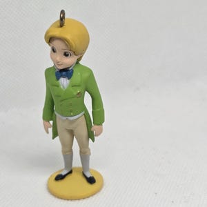 May include: A small figurine of a character with blonde hair, wearing a bright green tailcoat jacket, a blue bowtie, a white shirt, beige trousers, and grey socks with black shoes, standing on a round yellow base with a metal hanging loop.