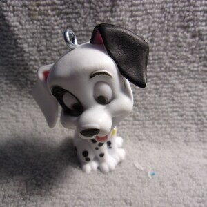 May include: A small, white Dalmatian puppy figurine with black spots, a black ear, and a silver loop for hanging. The puppy has a pink tongue and a yellow collar. The figurine is a collectible item.
