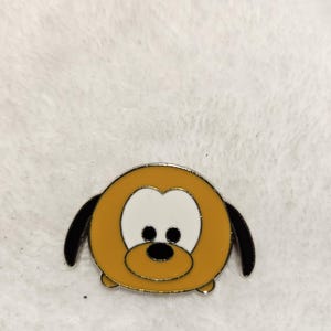 May include: A Pluto enamel pin featuring a rounded design with a yellow body, black ears, and a white face. The pin has a silver border and black eyes and nose. The pin is on a white, textured surface.