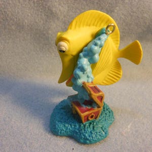 May include: A yellow fish figurine with a large dorsal fin and a metal hanging loop, positioned above a small red and gold treasure chest. A stream of light blue bubbles flows from the chest toward the fish, all set on a textured blue base.