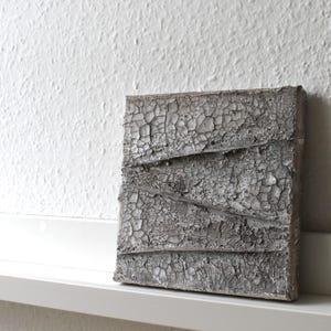 Könnte beinhalten: Ein quadratisches Wandkunstobjekt mit einer stark texturierten, rissigen grauen Oberfläche und drei markanten diagonalen Graten, die über die Vorderseite verlaufen. Das Finish wirkt verwittert und matt und erzeugt ein komplexes Muster aus Rissen.