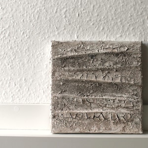 Puede incluir: Una obra de arte cuadrada y texturizada con apariencia desgastada. Presenta crestas horizontales en capas con un efecto agrietado y envejecido en tonos grises y beige. La obra se exhibe contra una pared blanca.