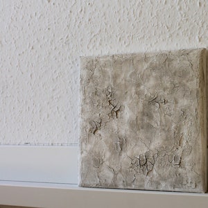 Könnte beinhalten: Ein kleines, quadratisches, graues Leinwandbild mit einer strukturierten Oberfläche. Die Struktur ähnelt rissiger Farbe oder einer verwitterten Oberfläche.