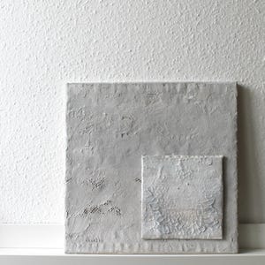 May include: A textured, square, light gray artwork with a smaller, attached square. The smaller square has a cracked, aged appearance. The background is a white, textured wall. The artwork is displayed on a white shelf.