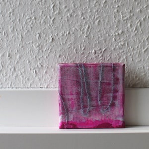 May include: A small pink and white canvas painting with a textured surface and gray lines. The painting is on a white shelf.