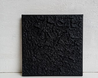Könnte beinhalten: Quadratische schwarze Leinwandkunst mit stark strukturierter Oberfläche. Das Kunstwerk ist einfarbig schwarz und hat eine raue, unebene Textur. Die Leinwand ist auf einem schwarzen Rahmen montiert. Der Hintergrund ist eine weiße Wand.
