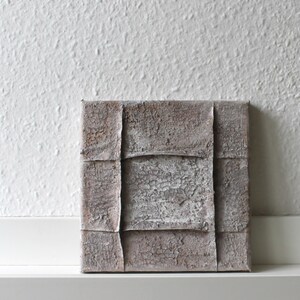 Puede incluir: Un cuadro cuadrado sobre lienzo con una superficie muy texturizada y agrietada en tonos topo, gris y blanco roto. La composición está dividida por dos crestas verticales y dos crestas horizontales en relieve, creando un patrón de cuadrícula.