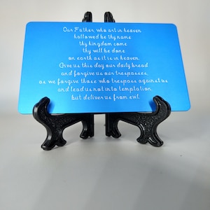 May include: A blue metal plaque with the Lord's Prayer engraved in white lettering. The plaque is displayed on a black metal stand.