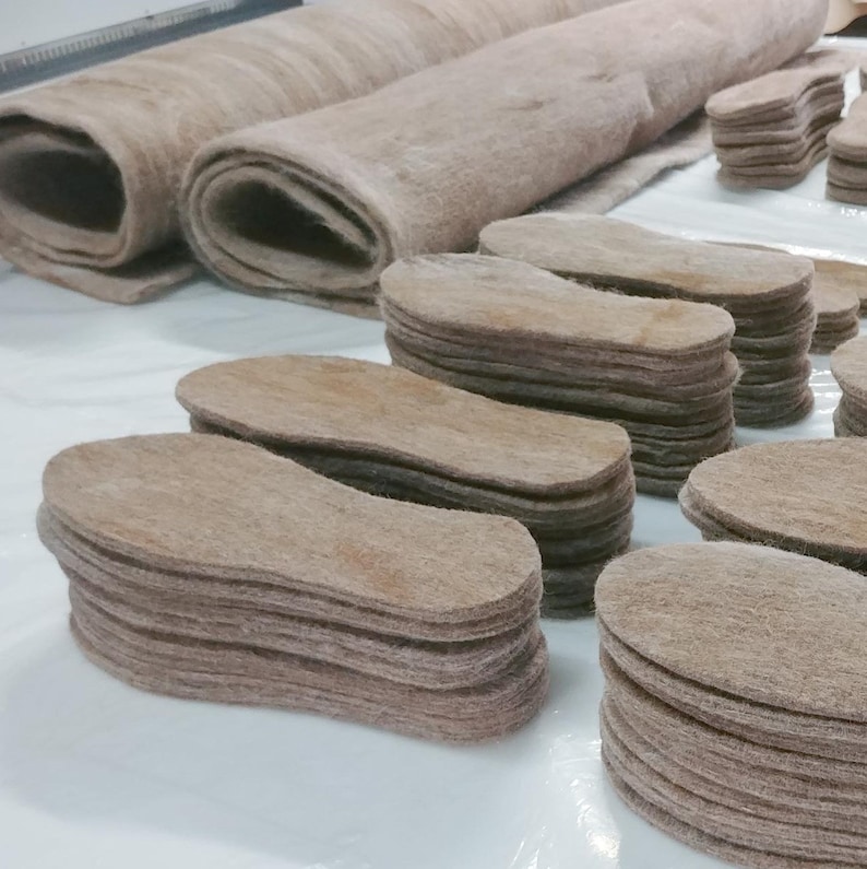 May include: A close-up of several stacks of brown felt insoles. The insoles are oval-shaped and are stacked neatly on a white surface.