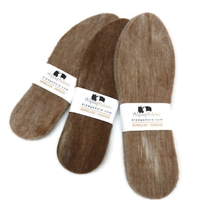 May include: Three brown alpaca wool insoles with white labels wrapped around them. The labels read "AlpagAdore" and "semelles - insoles". The insoles are oval-shaped and appear soft and fluffy.