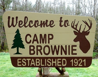May include: A wooden sign with the words "Welcome to Camp Brownie" in brown lettering. A green pine tree and a deer silhouette are also featured. The sign reads "Established 1921".