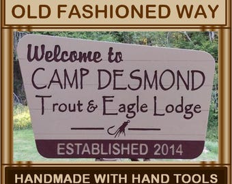May include: A rustic wooden sign with brown paint and white lettering welcomes visitors to Camp Desmond, a Trout & Eagle Lodge established in 2014. The sign is handmade with hand tools in Solitude Valley.