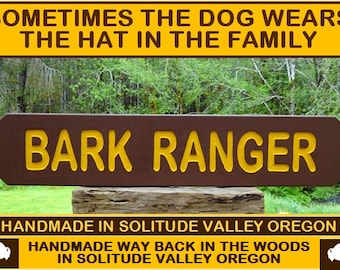 May include: A brown wooden sign with the text "BARK RANGER" in yellow letters. The sign is handmade in Solitude Valley, Oregon. The sign is in front of a green forest background.