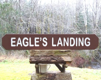 May include: A brown wooden sign with white lettering that reads "EAGLE'S LANDING." The sign is mounted on a rustic wooden structure with a small mug. The background features a forest and a stream.