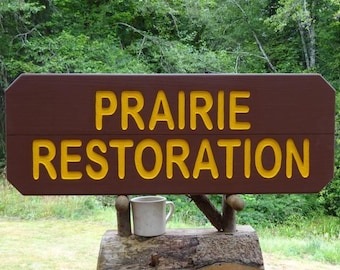 May include: A brown wooden sign with yellow lettering that reads "Prairie Restoration". The sign is mounted on a wooden post with a white ceramic mug sitting on top of the post.