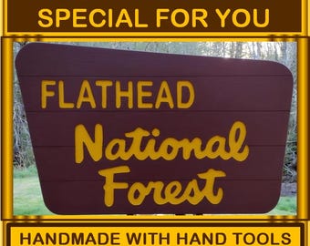 May include: A brown wooden sign with yellow lettering that reads "FLATHEAD National Forest." The sign also includes the text "WE CAN MAKE ONE SPECIAL FOR YOU" and "HANDMADE WITH HAND TOOLS IN SOLITUDE VALLEY OREGON."