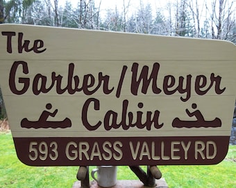 May include: A brown wooden sign with white lettering that reads "The Garber/Meyer Cabin 593 GRASS VALLEY RD". The sign has two brown canoe silhouettes with people paddling in them.