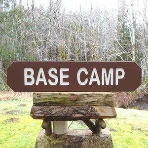 May include: A brown wooden sign with white lettering that reads "BASE CAMP". The sign is mounted on a rustic wooden post.