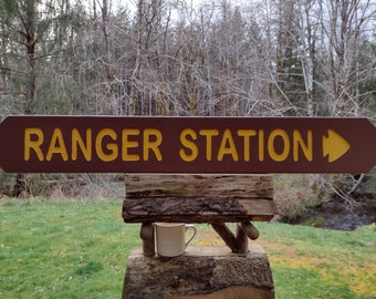 May include: A brown wooden sign with yellow text that reads "RANGER STATION" with a yellow arrow pointing to the right.