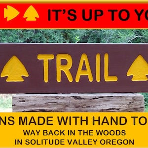 May include: A brown wooden sign with yellow text that reads "TRAIL" with yellow arrow symbols on either side. The sign is mounted on a weathered wooden post. The sign is surrounded by green trees. The text below the sign reads "SIGNS MADE WITH HAND TOOLS WAY BACK IN THE WOODS IN SOLITUDE VALLEY OREGON".