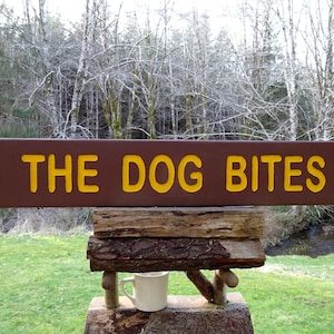 May include: A brown wooden sign with yellow lettering that reads "THE DOG BITES" is mounted on a rustic wooden structure. A white mug sits below the sign. The background features a grassy area and trees.