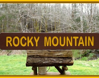 May include: A brown wooden sign with yellow lettering that reads "ROCKY MOUNTAIN". The sign is mounted on a wooden post and is in front of a forest.