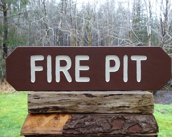 Puede incluir: Un letrero de madera marrón con letras blancas que dice "FIRE PIT". El letrero está montado en una pila de troncos de madera desgastados.
