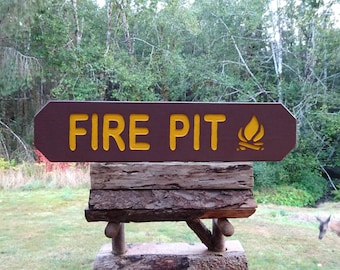 May include: A brown wooden sign with the words "FIRE PIT" in yellow, and a flame icon. The sign is mounted on a rustic wooden base, with a natural green background of trees and grass.