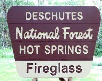 May include: A brown and white sign with the text "Deschutes National Forest Hot Springs Fireglass" in white lettering.