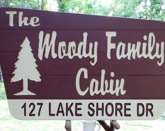 May include: A wooden sign with a maroon background and white lettering. The sign reads "The Moody Family Cabin" with a tree graphic. The bottom section of the sign is white and displays "127 LAKE SHORE DR".