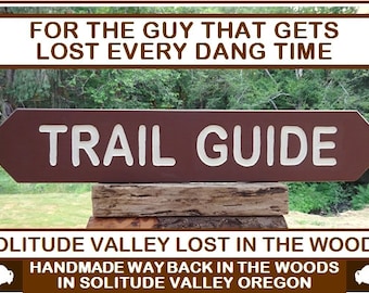 May include: A brown wooden sign with white lettering that reads "TRAIL GUIDE" and the text "FOR THE GUY THAT GETS LOST EVERY DANG TIME" above it. The sign is mounted on a wooden post and is surrounded by green foliage. The text below the sign reads "SOLITUDE VALLEY LOST IN THE WOODS HANDMADE WAY BACK IN THE WOODS IN SOLITUDE VALLEY OREGON".