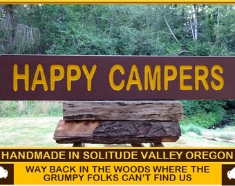 May include: A brown wooden sign with the text "HAPPY CAMPERS" in yellow letters. The sign is sitting on a stack of wooden logs. The text below the sign reads "HANDMADE IN SOLITUDE VALLEY OREGON" and "WAY BACK IN THE WOODS WHERE THE GRUMPY FOLKS CAN'T FIND US".