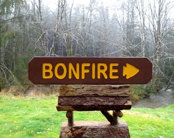 May include: A brown wooden sign with yellow text that reads "BONFIRE" and a yellow arrow pointing to the right. The sign is mounted on a wooden post in a forest setting.