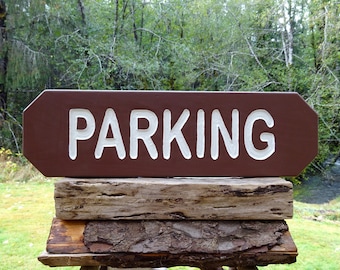 Puede incluir: Un letrero de madera marrón con bordes redondeados y letras blancas que dice "PARKING". El letrero está colocado sobre una pila de troncos de madera desgastados.