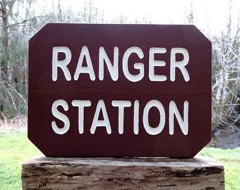 Puede incluir: Un letrero rectangular marrón con las palabras "RANGER STATION" en blanco. El letrero está montado sobre un poste de madera. El fondo muestra un bosque y un arroyo.