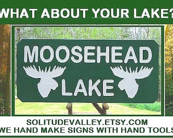 May include: A green rectangular sign with white lettering that reads "MOOSEHEAD LAKE" with moose head silhouettes. The sign also includes the text "WHAT ABOUT YOUR LAKE?" and "SOLITUDEVALLEY.ETSY.COM".