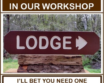 May include: A handcrafted wooden sign with the word "LODGE" in white lettering and an arrow. The sign is dark red-brown and mounted on a rustic wooden base. The text "WE MAKE IT FRESH IN OUR WORKSHOP" and "I'LL BET YOU NEED ONE FOR YOUR LODGE" are also displayed.