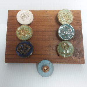 May include: A set of seven ceramic knobs with various ocean-themed designs, including a seashell, a fish, a turtle, a palm tree, and a sailboat. The knobs are displayed on a wooden board.