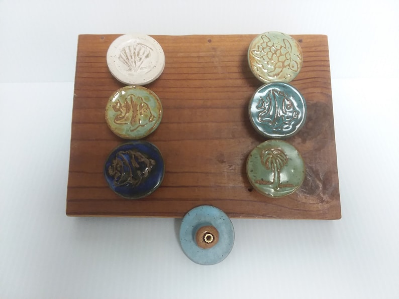 Może przedstawiać: Zestaw siedmiu ceramicznych gałek z r&oacute;żnymi wzorami o tematyce oceanicznej, w tym muszla, ryba, ż&oacute;łw, palma i żagl&oacute;wka. Gałki są prezentowane na drewnianej desce.