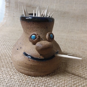May include: A brown ceramic toothpick holder shaped like a face with blue eyes and a black smile. The holder has a black rim and is filled with toothpicks.