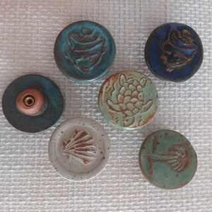 Może przedstawiać: Sześć ceramicznych gałek z r&oacute;żnymi wzorami o tematyce oceanicznej. Gałki są okrągłe i mają wypukły wz&oacute;r. Wzory obejmują rybę, ż&oacute;łwia, muszlę, palmę i dwie r&oacute;żne ryby.