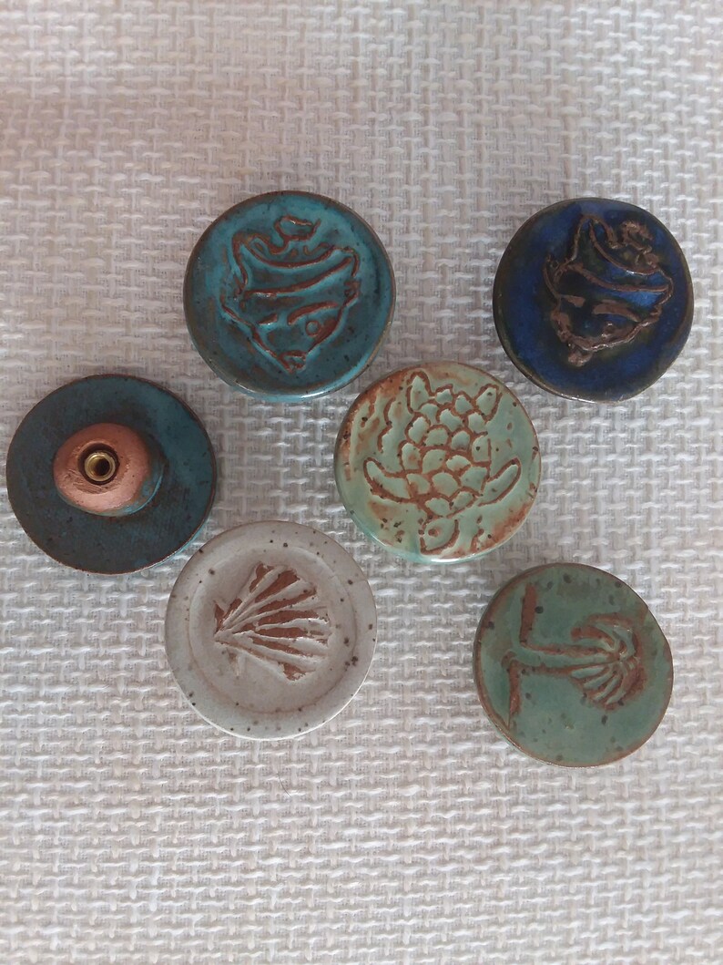 Może przedstawiać: Sześć ceramicznych gałek z r&oacute;żnymi wzorami o tematyce oceanicznej. Gałki są okrągłe i mają wypukły wz&oacute;r. Wzory obejmują rybę, ż&oacute;łwia, muszlę, palmę i dwie r&oacute;żne ryby.