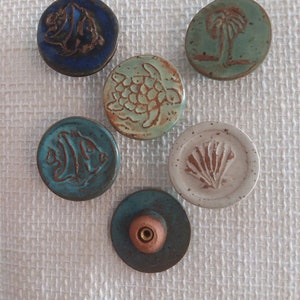 Może przedstawiać: Sześć okrągłych ozd&oacute;b ceramicznych z r&oacute;żnymi wzorami o tematyce oceanicznej. Ozdoby są niebieskie, zielone, białe i brązowe. Każda ozdoba ma inny wz&oacute;r, w tym rybę, ż&oacute;łwia, muszlę, palmę i rybę.