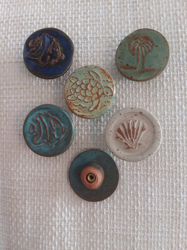 Może przedstawiać: Sześć okrągłych ozd&oacute;b ceramicznych z r&oacute;żnymi wzorami o tematyce oceanicznej. Ozdoby są niebieskie, zielone, białe i brązowe. Każda ozdoba ma inny wz&oacute;r, w tym rybę, ż&oacute;łwia, muszlę, palmę i rybę.