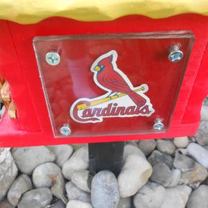 May include: A red and yellow wooden birdhouse with a clear plastic panel displaying the St. Louis Cardinals logo. The birdhouse is mounted on a black post and surrounded by rocks.