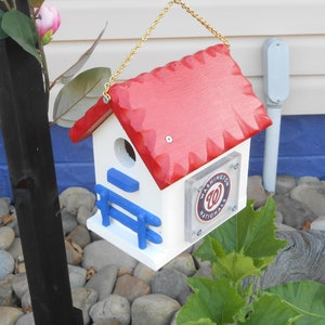 May include: A white birdhouse with a red roof and a blue porch. The birdhouse has a clear plastic panel on the side with the Washington Nationals logo. The birdhouse is hanging from a gold chain.