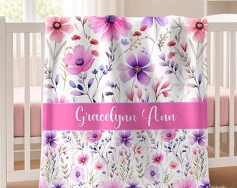 May include: A white baby blanket featuring a watercolor floral pattern in shades of pink and purple. A horizontal pink band across the center displays the name Gracelynn Ann in white cursive script. The blanket is draped over a white crib.