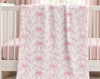 May include: A white blanket featuring a repeating pattern of pink bows, floral sprigs, and the name Ava Leigh printed in a cursive script. The design is consistent across the fabric, showcasing soft pink illustrations on a clean white background.